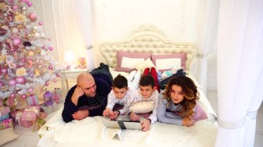 Uyumlu aile eğlenceli ve birlikte saat karikatürler bilgisayarınızda sahip, Noel ağacı ve hediyeler ile parlak yatak yatakta yatarken.