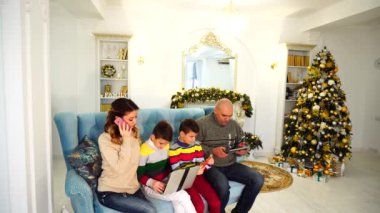 Modern ve mobil aile, iki oğlu ve karı kendi işleri ile meşgul ve Noel ağacı ile şenlikli bir şekilde dekore edilmiş oda mavi kanepede oturan eğlence için araçları kullanmak ve