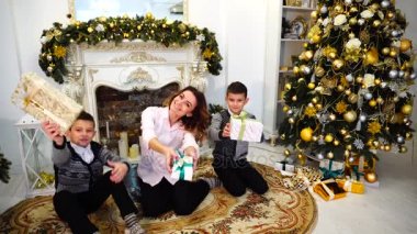 Güzel kadın ve anne ve çocuk çocuklar göz ve kamera iyi ruh hali ve katta oturma odasında oturup ellerinde hediyelerle gülümseme ile dekore edilmiş şömine ve yüksek çam ağacı ile tatil için.