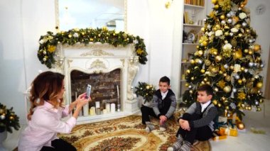Anne oğlu oğlu aile albümü için smartphone çekiyor ve oturma odasında şömine ve yüksek çam ağacı ile tatil için dekore edilmiş katta oturur.