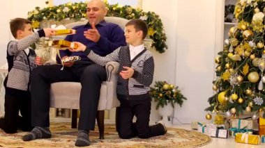 Aile babası ve oğulları gülümser ve birbirlerine hediyeler şömine ve yeni yılda dekore edilmiş uzun Noel ağacı ile oturma odasında sakso.