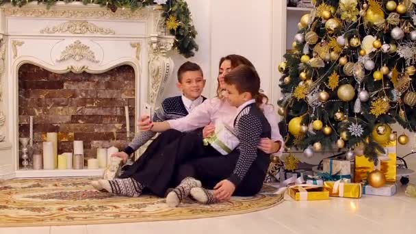 Maravillosa hembra, madre de dos hijos hace selfie en el teléfono inteligente con gemelos, sentado en el suelo en la sala de estar, decorado para vacaciones con chimenea y abeto alto . — Vídeos de Stock