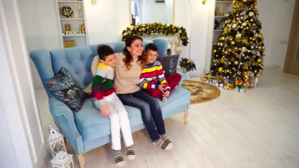 Feliz madre de dos hijos y hermanos niños se sientan lado a lado y en el camino de la familia abrazan en el sofá azul en la habitación decorada festiva con árbol de Navidad y chimenea en el día . — Vídeo de stock