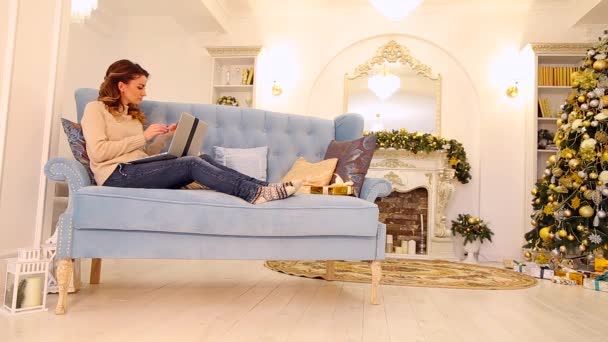 Mère commande des cadeaux sur Internet sur ordinateur pour les fils et paie une carte en plastique, assise sur un canapé bleu dans une chambre décorée avec un arbre de Noël et une cheminée pendant la journée . — Video