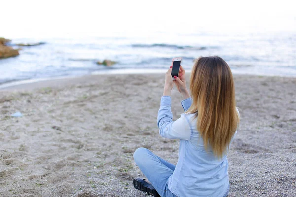 Chica en la orilla hace foto en el teléfono inteligente, sentado en la orilla arenosa — Foto de Stock