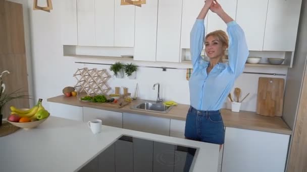 Menina bonita entra na cozinha e se estende, desfrutando de café da manhã, de pé no meio da cozinha na mesa da cozinha . — Vídeo de Stock