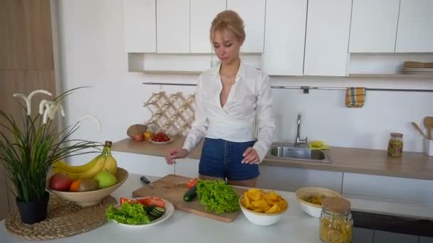 Slanke blonde vrouw voltooit voorbereiding van gezond ontbijt en verspreidt groenten, permanent aan tafel van moderne keuken. — Stockvideo