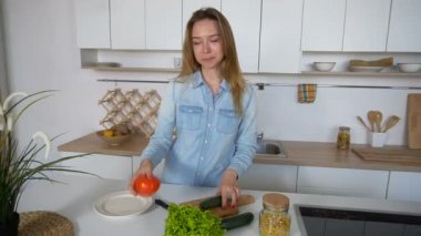 Modern kız kamera, mutfak, mutfak masasının ortasına sabah ayakta içine gülümser ve salata hazırlamak için hangi bitkisel seçer.