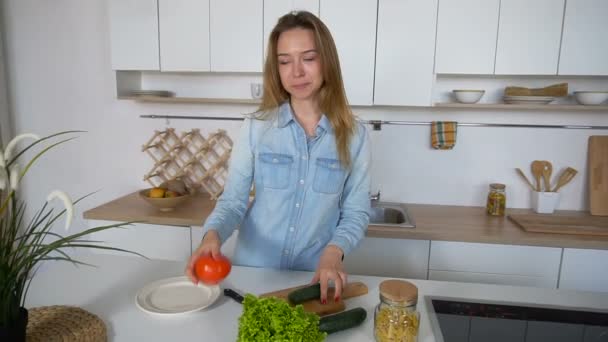 Nowoczesna dziewczyna wybiera z których warzyw przygotować sałatkę i uśmiecha się do kamery, stojąc w środku kuchni przy stole w kuchni rano. — Wideo stockowe