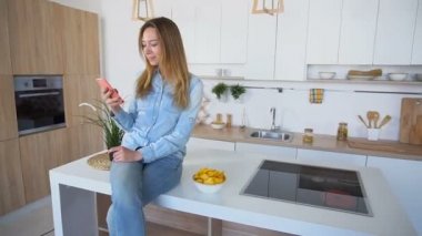 Güzel kadın cep telefonu yazışmalar için kullanır ve patates cipsi mutfak masa yüzeyine oturan çalışır.