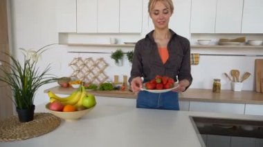 Güzel kadın poz ve kamera modern mutfakta olgun çilek plaka ile gülümseyen.