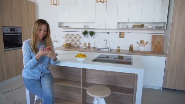 Chica atractiva sosteniendo el teléfono y comer papas fritas sentado en la silla en la mesa de la cocina . — Vídeo de stock
