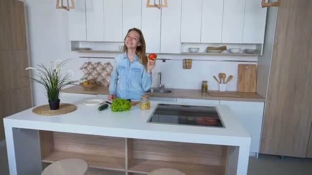 Dona de casa alegre sorrindo com tomate nas mãos e preparando salada, de pé no meio da cozinha durante o dia . — Vídeo de Stock