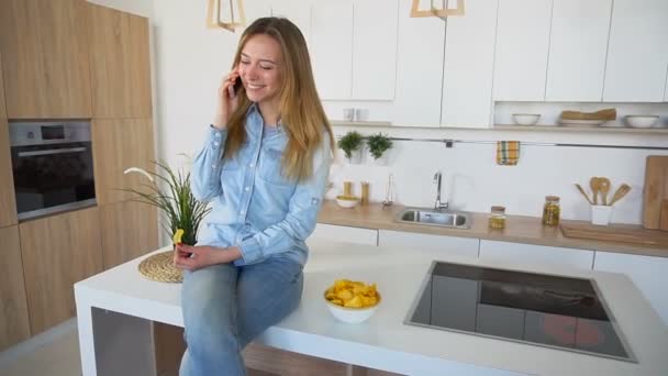 Schöne junge Frau plaudert am Telefon und isst Kartoffelchips, die auf der Oberfläche des Küchentisches sitzen. — Stockvideo