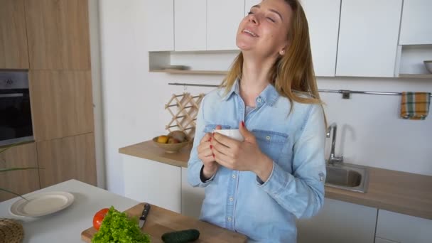 Giovane donna felice di iniziare un nuovo giorno con una tazza di tè in mano, in piedi in mezzo alla cucina al tavolo della cucina al mattino . — Video Stock