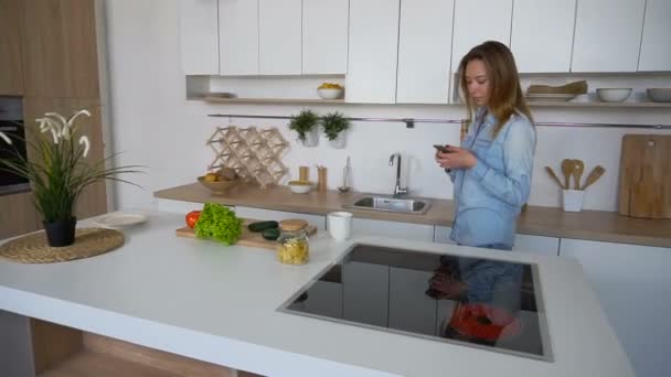 Biznes Kobieta wchodzi kuchnia z telefonem w ręce i bierze łyk herbaty, stojąc w środku kuchni przy stole w kuchni rano. — Wideo stockowe