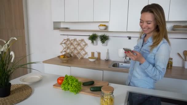 Κομψό κορίτσι χρησιμοποιεί κινητό και κρατά την κούπα του τσαγιού στα χέρια, στέκεται στη μέση του κουζίνα στο τραπέζι της κουζίνας πρωί. — Αρχείο Βίντεο