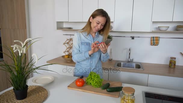 現代の少女、日中にスタイリッシュなキッチンの真ん中に立って、社会的なネットワークのためにスマート フォン上の野菜の写真. — ストック動画