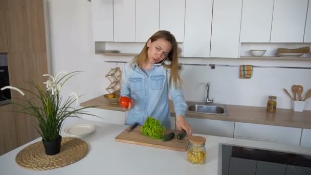 Příjemná žena diskutujících na buňku a jde připravit salát ze zeleniny, ve středu stylové kuchyně v odpoledne. — Stock video