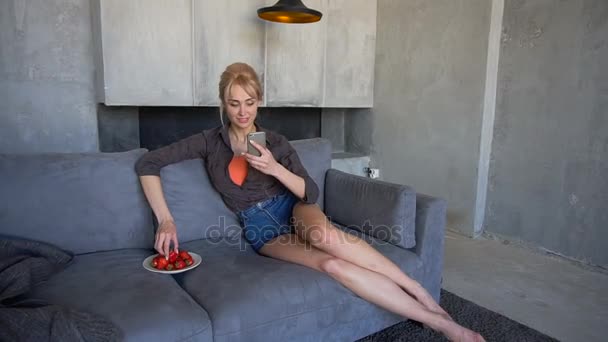 Blond woman holds mobile phone and eats ripe strawberry, sitting on soft sofa in living room with gray walls in afternoon. — Stock Video