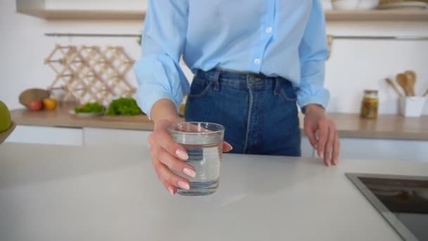 Hermosa mujer sacia la sed con un vaso de agua y se encuentra en medio de la cocina en la mesa de la cocina durante el día . — Vídeo de stock