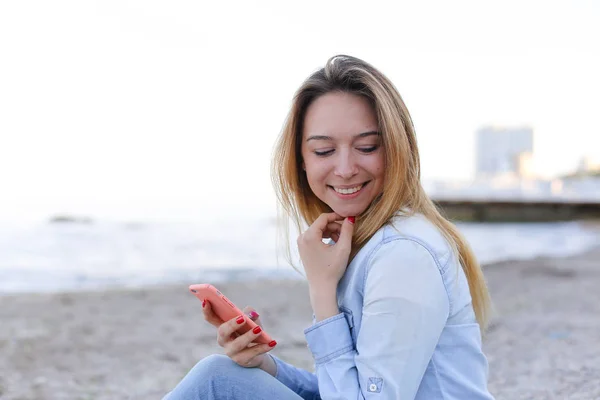 Mooi meisje chatters op telefoon met glimlach en zit op strand n — Stockfoto