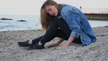 Gülümseyen genç kadın kumsalda aittir ve kamera, kumsalda kabukları ile sıcak bahar akşamı oturan pozlar.