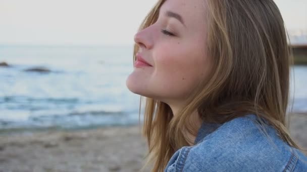 Slow Motion Portrait of cheerful girl who poses on camera and cute laughs sitting on sand on background of blue sea and sky at sunset in summer. — Stock Video