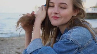 Kim kamera ve sevimli yaz aylarında gün batımında mavi deniz ve gökyüzü karşı sahilde otururken gülerek poz neşeli kız portresi hareket yavaş.
