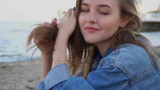 Lassú mozgás vidám lány portréja, aki pózol a kamera, és aranyos nevetve, miközben ül a strandon, szemben a kék tenger és az ég naplemente a nyári időszakban. — Stock videók
