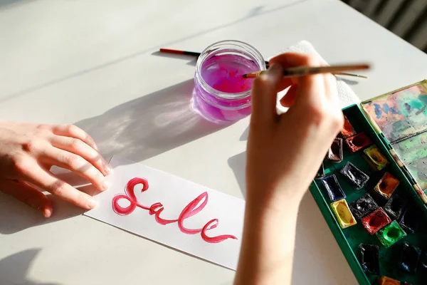 Chica pintor escribe con pincel y pinturas inscripción en hoja , — Foto de Stock