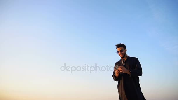 Homem moderno árabe viajante detém smartphone e tira fotos de si mesmo como lembrança, de pé no meio do deserto arenoso ao ar livre na noite de verão . — Vídeo de Stock