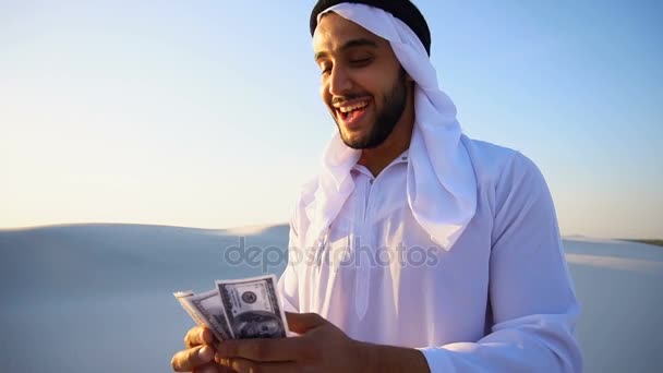 El sheikh exitoso árabe considera los billetes y feliz, de pie en el centro del desierto arenoso en el día caliente veraniego . — Vídeo de stock