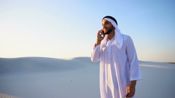 Muslimska manliga affärsman pratar på telefon och delar nyheter med följeslagare, stående i mitten av sandig öken varm sommarkväll. — Stockvideo