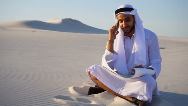 Portrait of Muslim designer Arabian Sheikh who coordinates on mobile with client building and sits on sand with project in wide desert on hot morning. — Stock Video