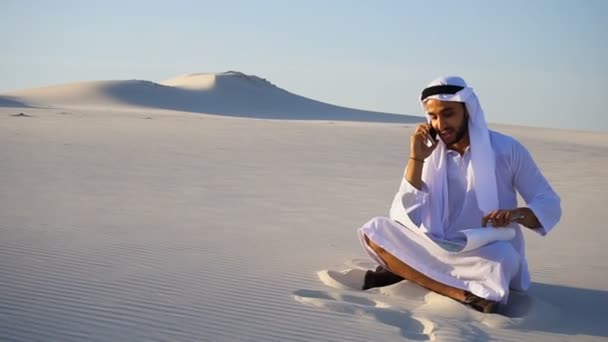 Vertrouwen mannelijke Arabische Uae Sheikh bouwer spreekt op mobiele met klant, zittend op zand met papier tekenen in grote woestijn op warme dag. — Stockvideo