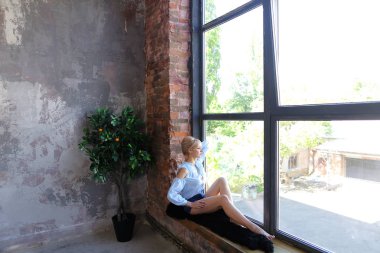 Çekici genç kadın gülümseyerek poz veriyor ve pencere pervazına üzerinde oturur