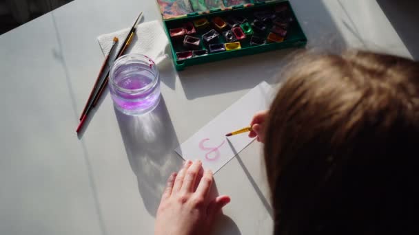 Chica pintor escribe con pincel y pinturas inscripción en la hoja, sentado a la mesa con materiales de arte en el taller durante el día . — Vídeos de Stock