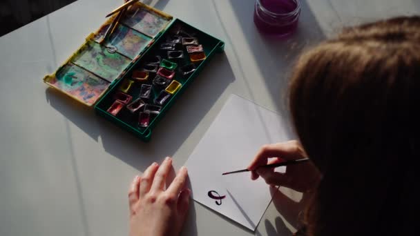 La artista en cursiva escribe la inscripción en cursiva en la hoja de papel y se sienta a la mesa con materiales de arte en el taller por la tarde . — Vídeos de Stock
