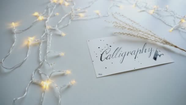 Close-up tiro de cartão postal com itálico caligrafia lettering em folha de papel branco entre guirlanda na mesa de luz . — Vídeo de Stock
