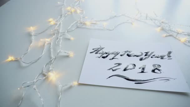 Carte de vœux festive avec nouvelle année faite à l'encre noire sur papier feuille blanche sur table avec guirlande . — Video