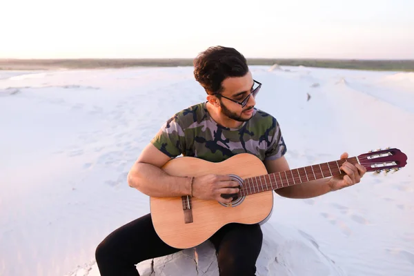 Cute young male Arab enjoys sound of guitar, sitting on hill in