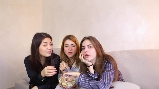 Amichevoli amiche guardano film e mangiano popcorn, seduti su un morbido divano in una luminosa camera da letto la sera . — Video Stock