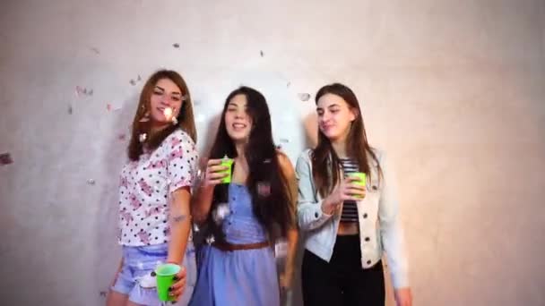 Funny girls dance and drink american beer in camera with smiles on their faces and stand against background of light wall in room. — Stock Video