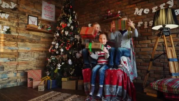 Küçük çocuklar kamera, koltukta oturan hediyeler vermek mutlu kardeşler şenlikli odaya dekore edilmiş — Stok video