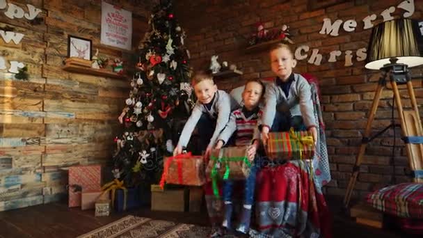 Trzech chłopców, dzieci zachować nowy rok prezenty w swoje ręce i wstrząsnąć z ciekawości, siedząc na fotelu urządzony pokój uroczysty — Wideo stockowe