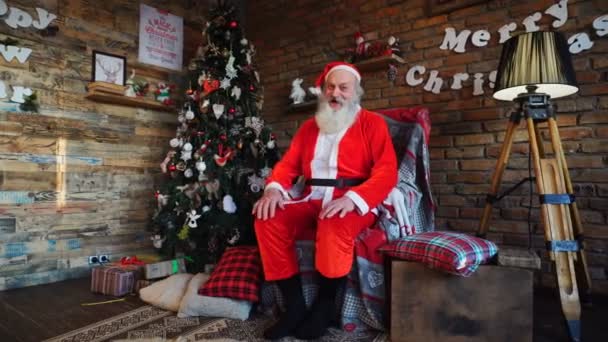 Energiczny Santa Claus podnosi kciuk, siedząc na fotelu w świątecznie urządzony pokój — Wideo stockowe