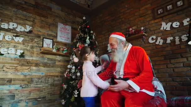 Kleines Mädchen umarmt Weihnachtsmann und wünscht sich Weihnachten in gemütlich dekoriertem Raum für den Urlaub — Stockvideo