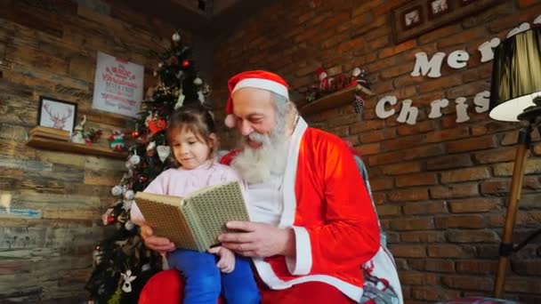 Santa Claus sitzt auf einem Sessel und liest im geschmückten Festsaal Märchen für begeisterte kleine Mädchen — Stockvideo