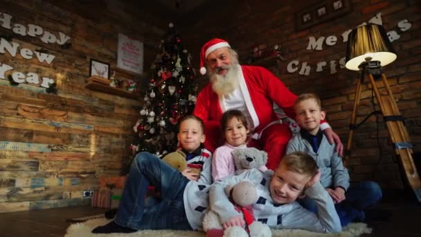 Дружелюбные дети и Санта-Клаус позируют на камеру в украшенной высокой елкой комнате — стоковое видео